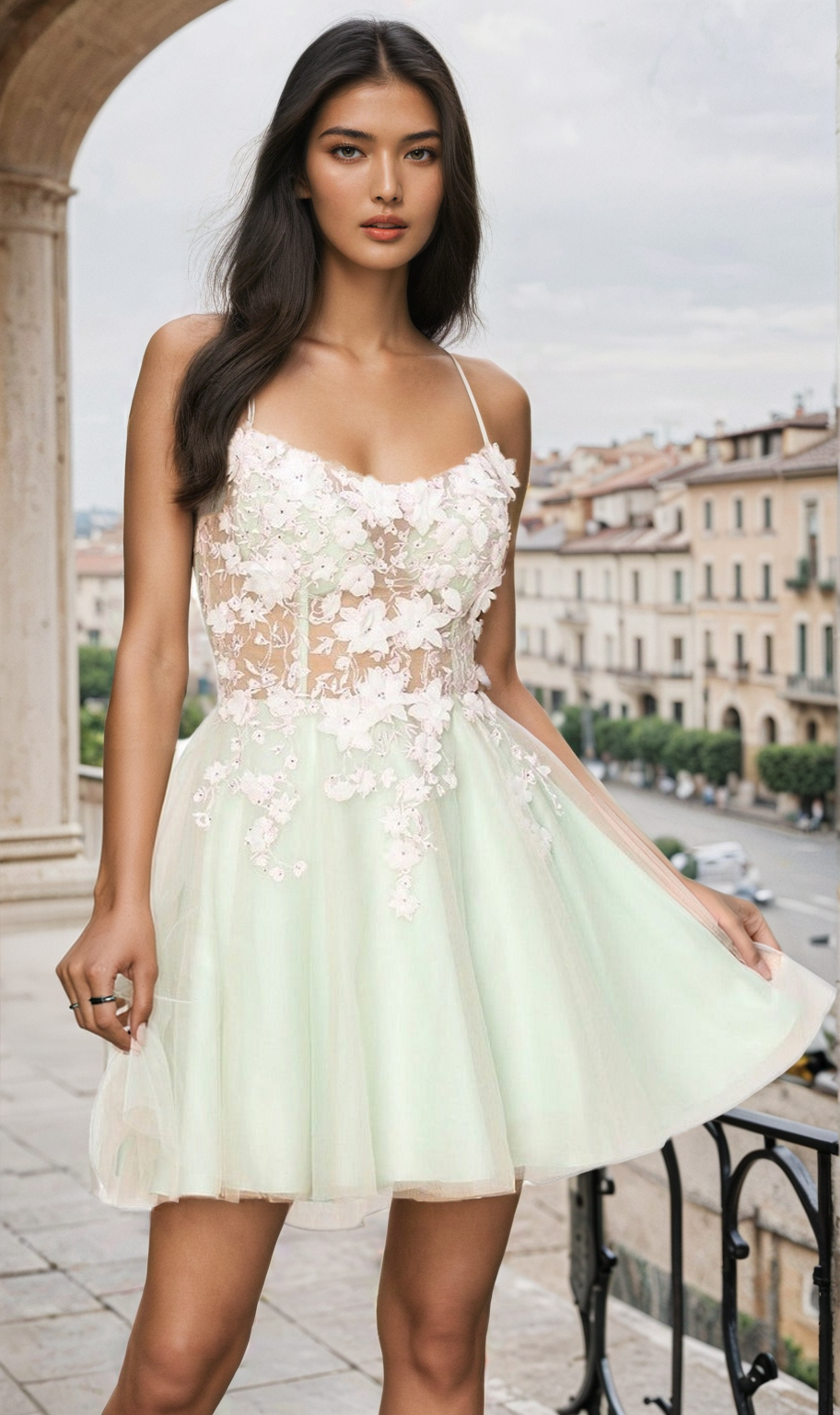 A woman stands on a balcony wearing the Backless Sheer-Waist Floral Homecoming Dress 8053J—a light green short dress with 3-D flowers and sheer details—looking at the camera. Buildings and a street are visible in the background.